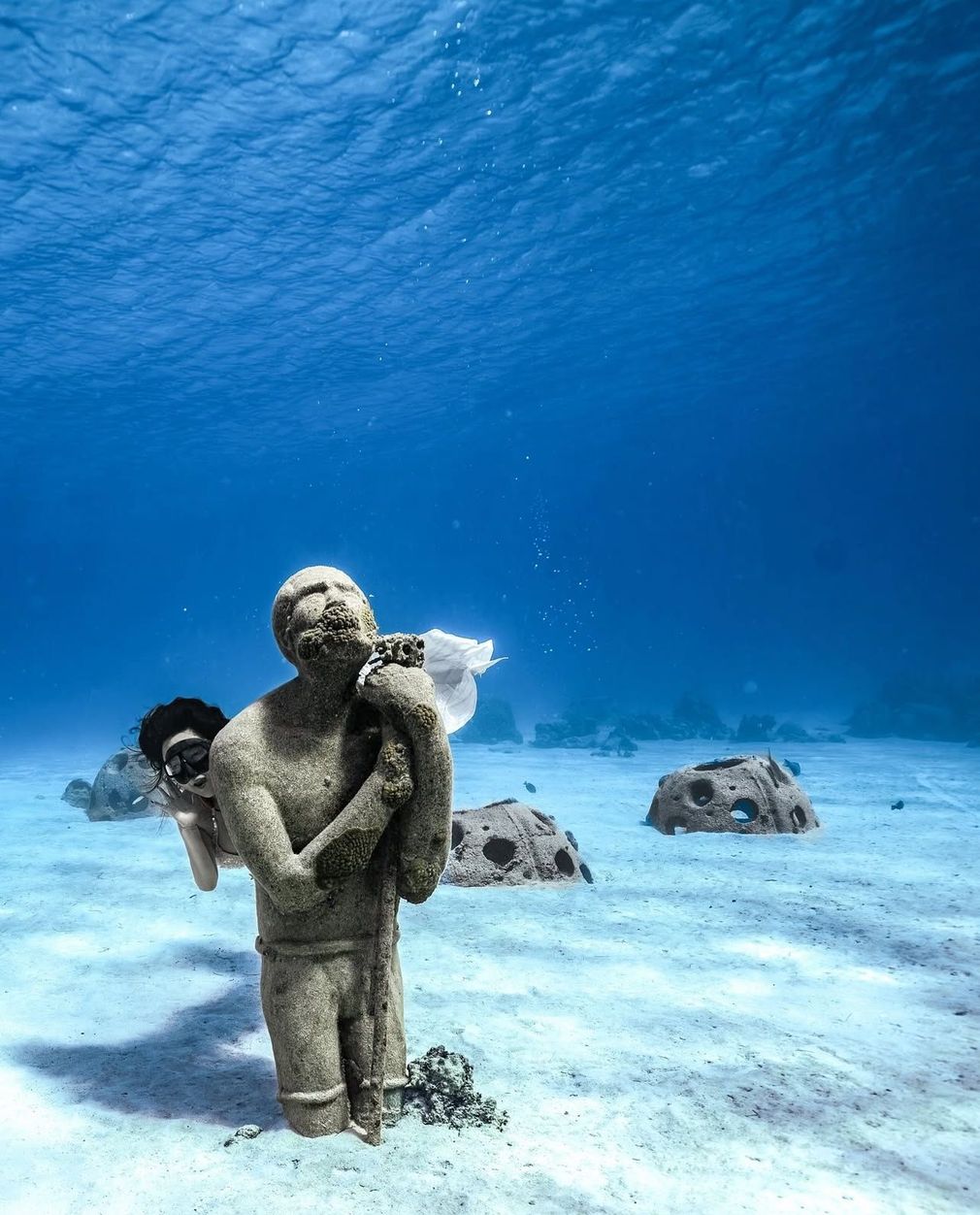 Underwater statue of a person carrying another, on a sandy seabed, with blue water above.