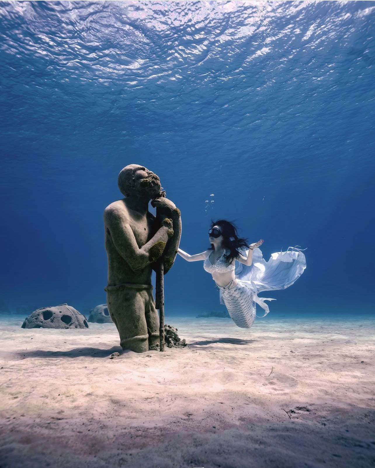 Underwater sculpture and person in flowing white dress, posing near sandy ocean floor.