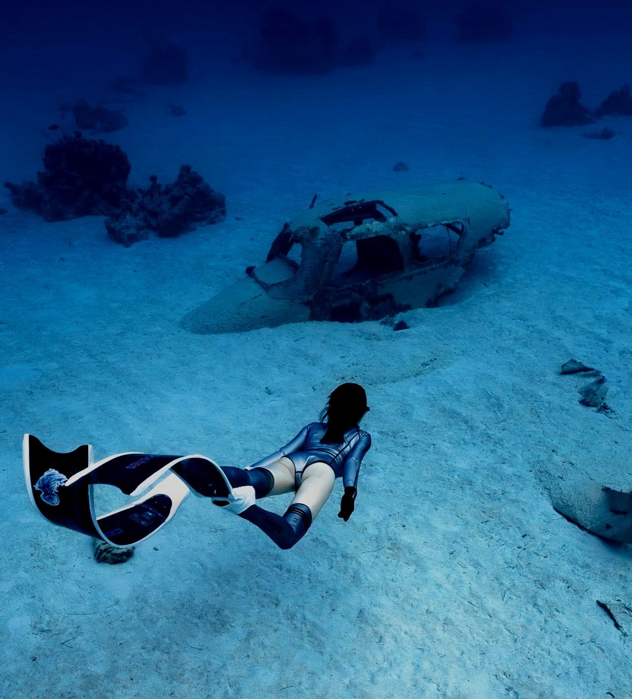Freediver swims past a submerged plane, sandy seabed, dark blue water.