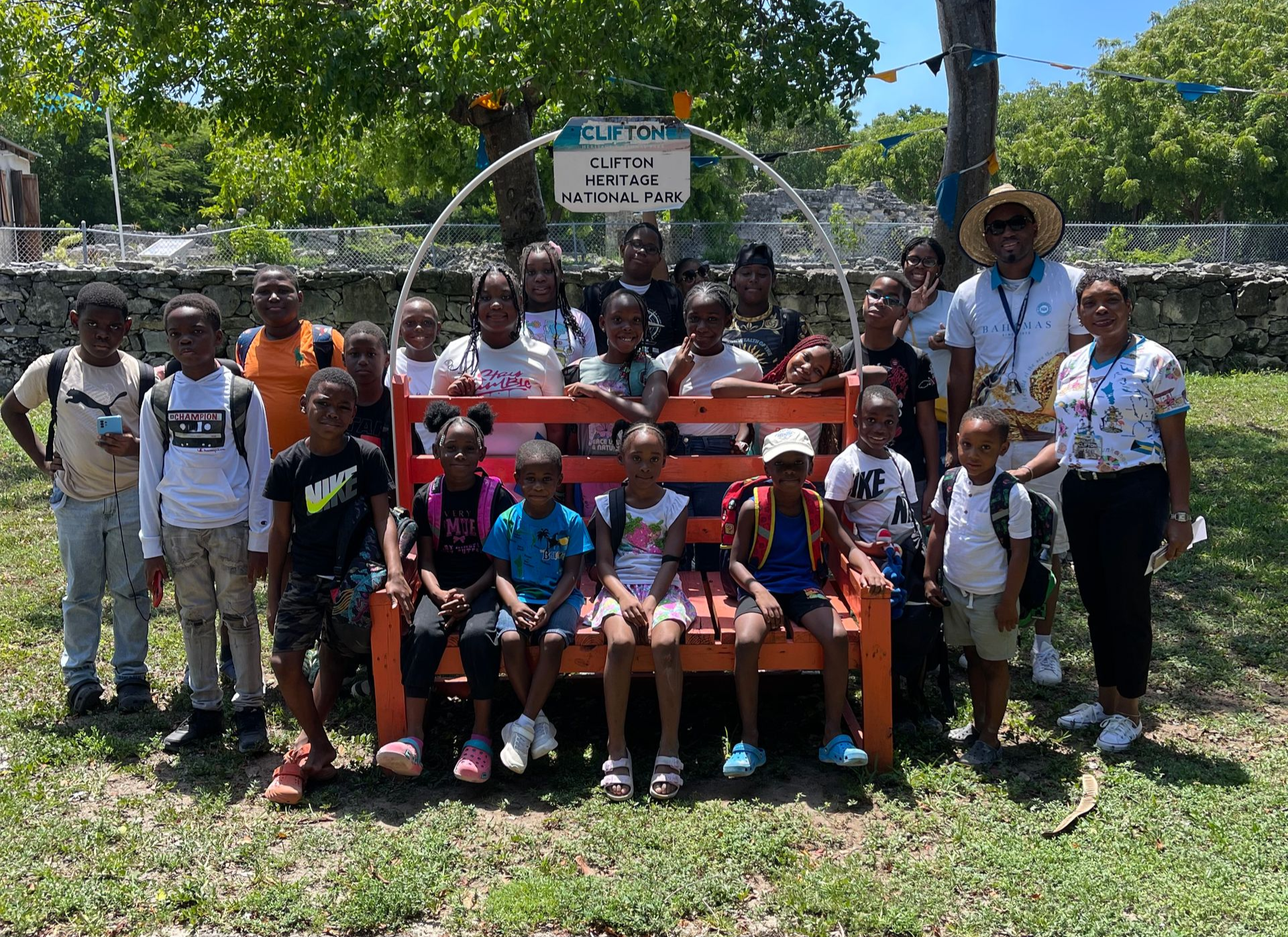 Group of children on a large orange bench with adults in a sunny outdoor setting.