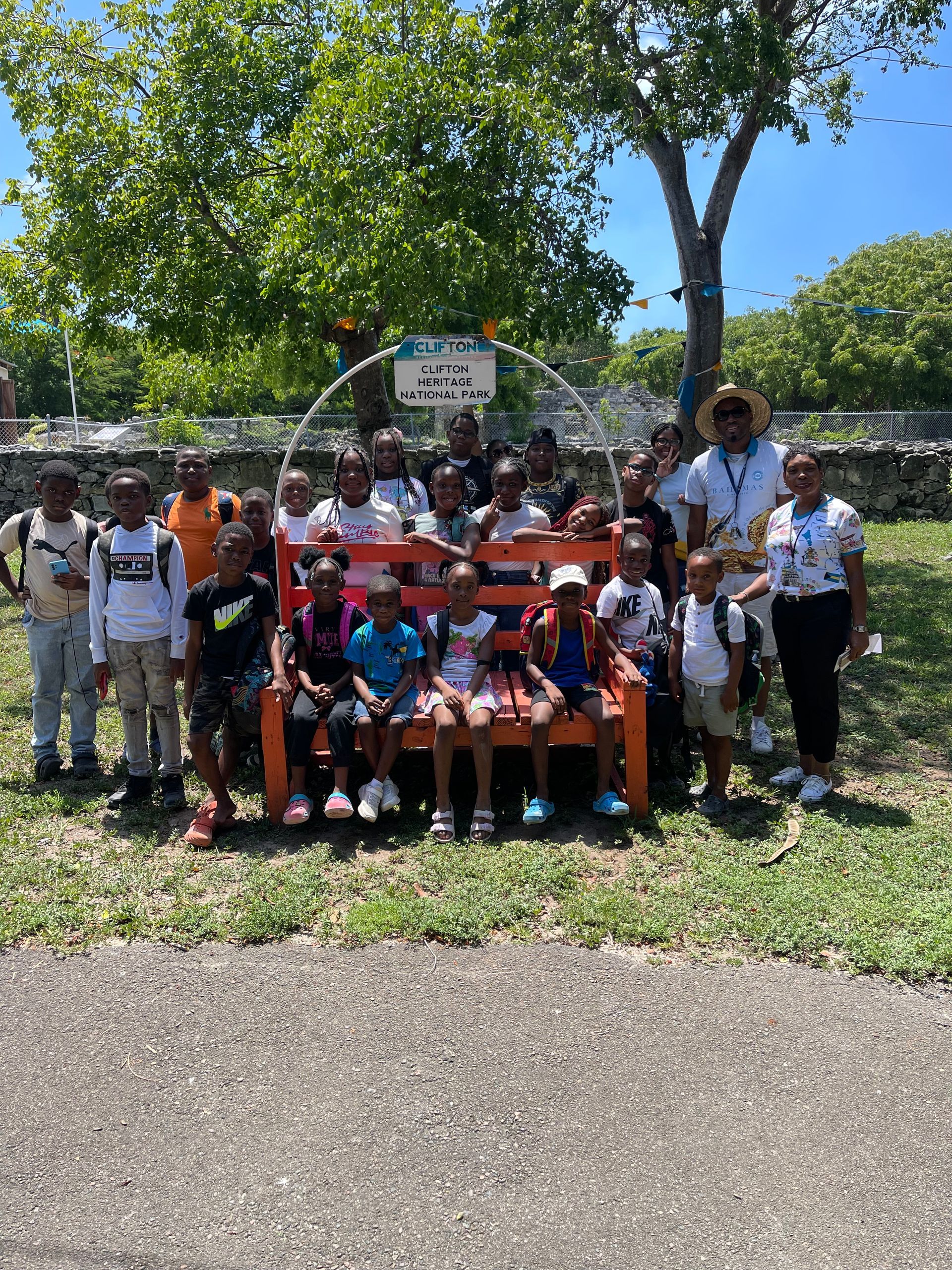 Group of children sitting and standing on and around an orange bench with two adults outdoors.