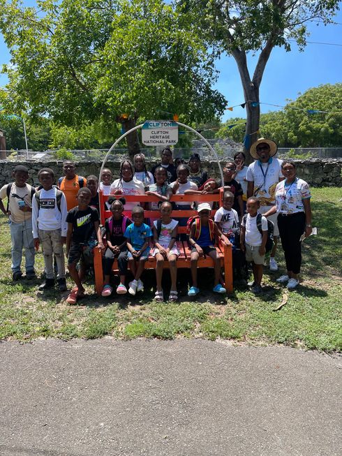Group of children posing with adults on a large, orange bench outdoors, under a sign.