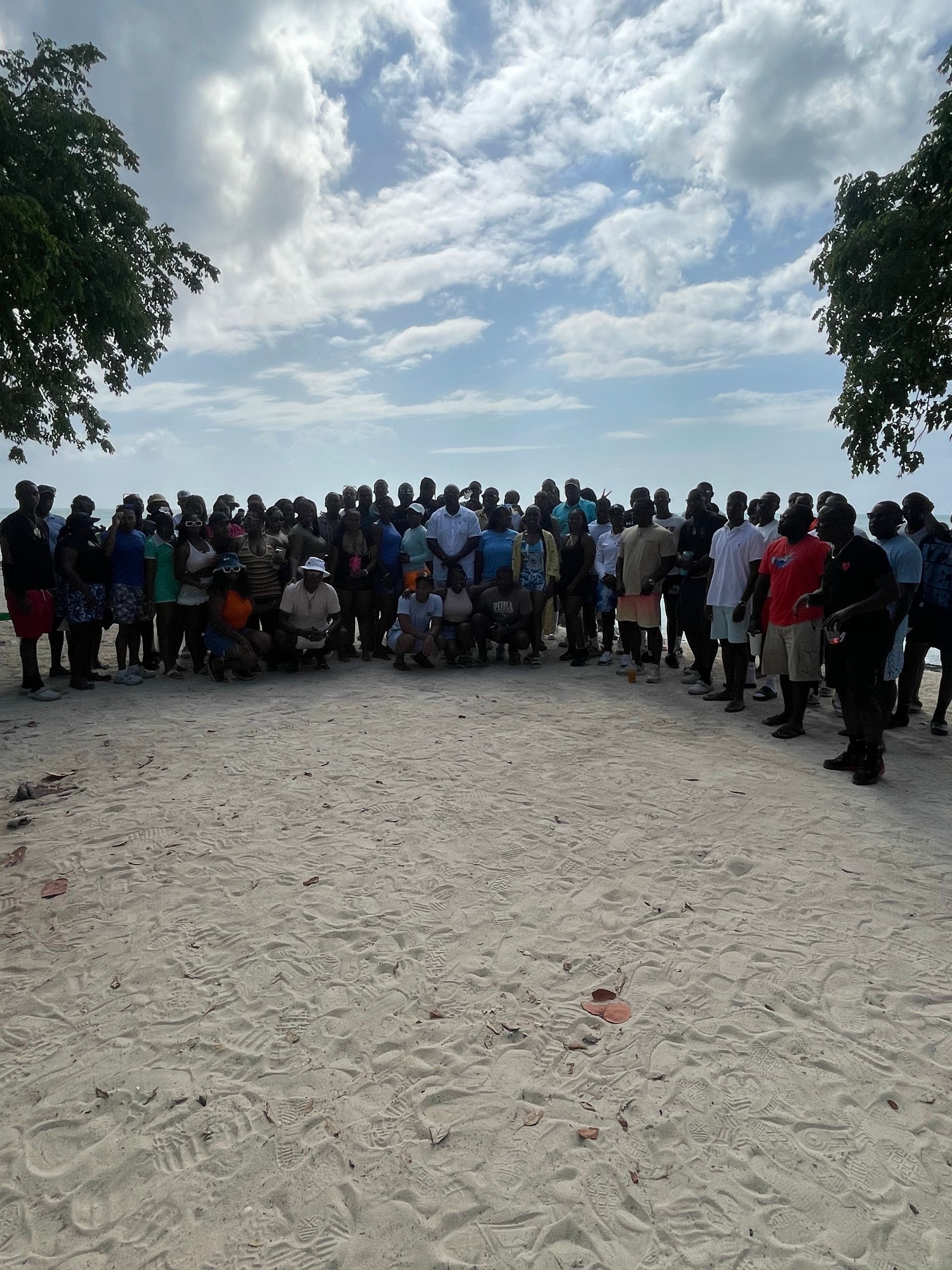 Large group of people on a sandy beach under a cloudy sky near trees.