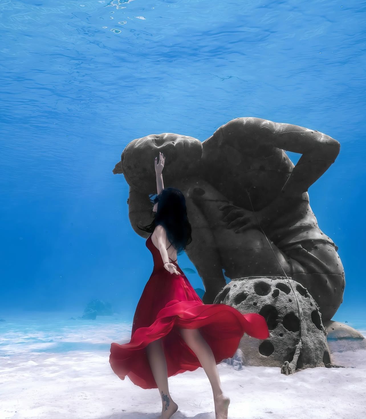 Woman in red dress underwater, reaching toward a large submerged statue.