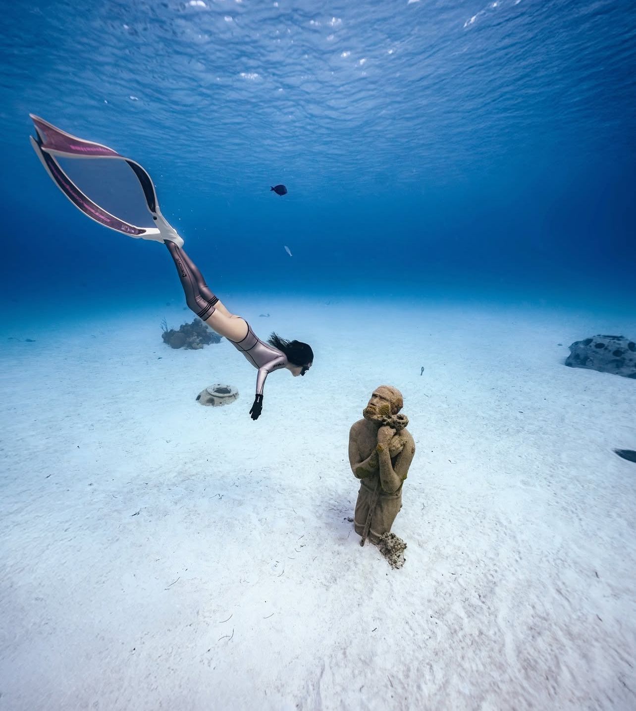 A person in a mermaid tail swims underwater near a statue on a sandy ocean floor.