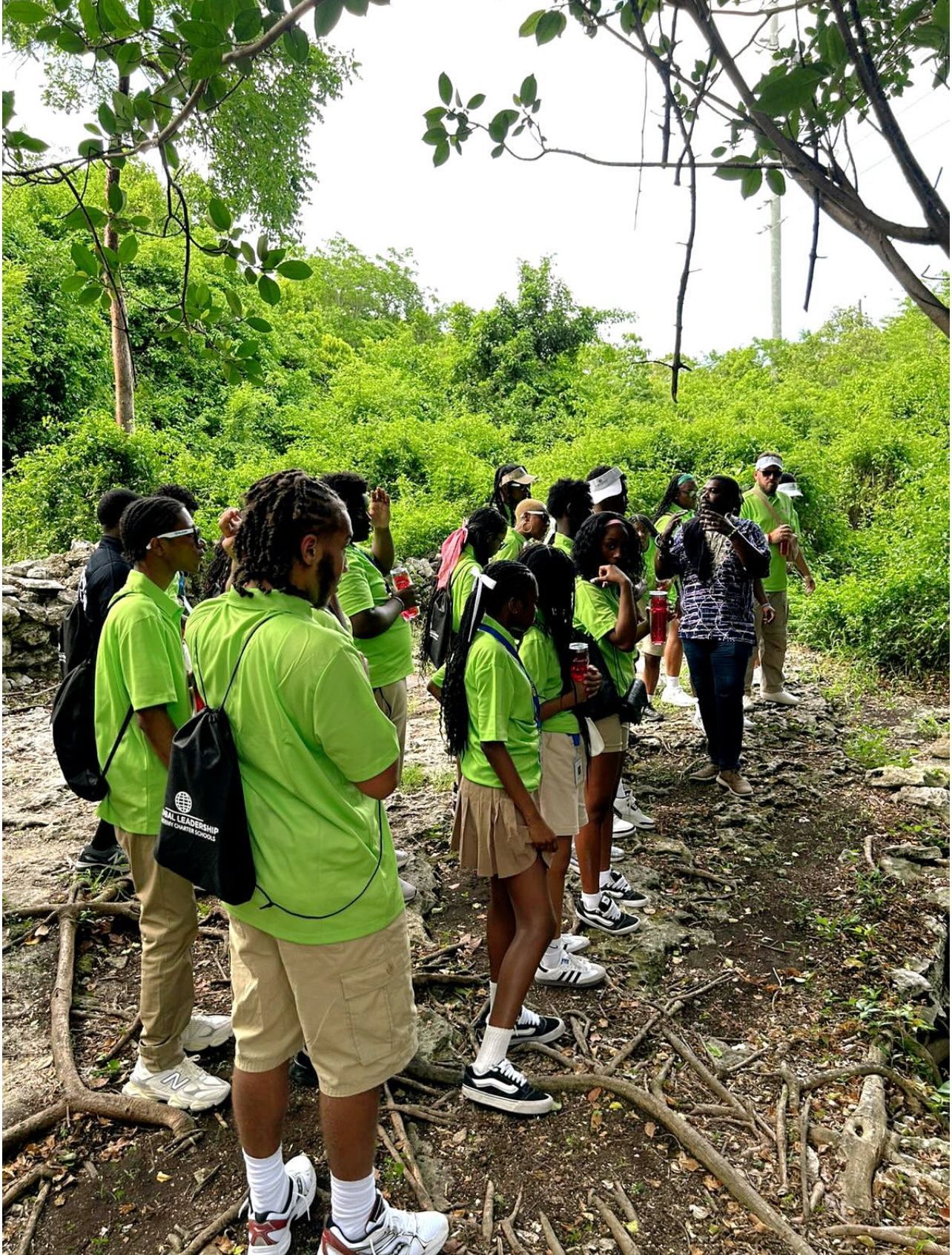 Group of people in green shirts on a wooded path, looking toward a person speaking.