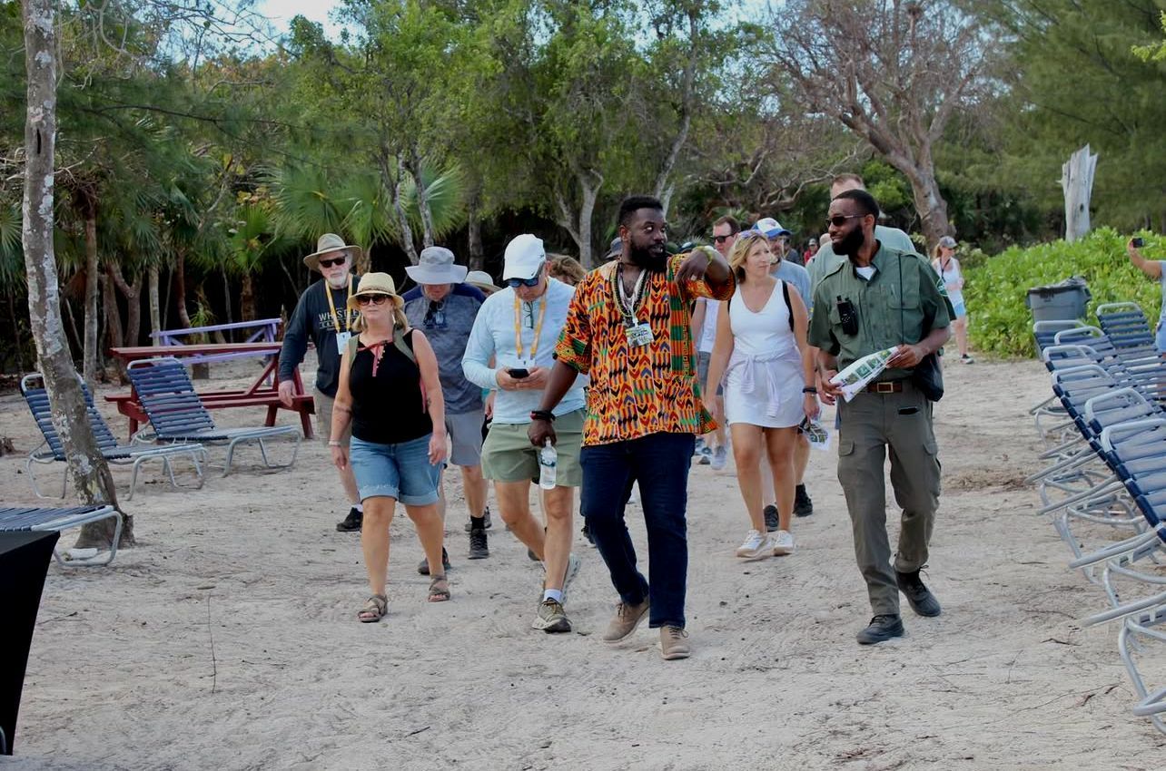 Group of people walking on sand near trees, led by two men, sunny day.
