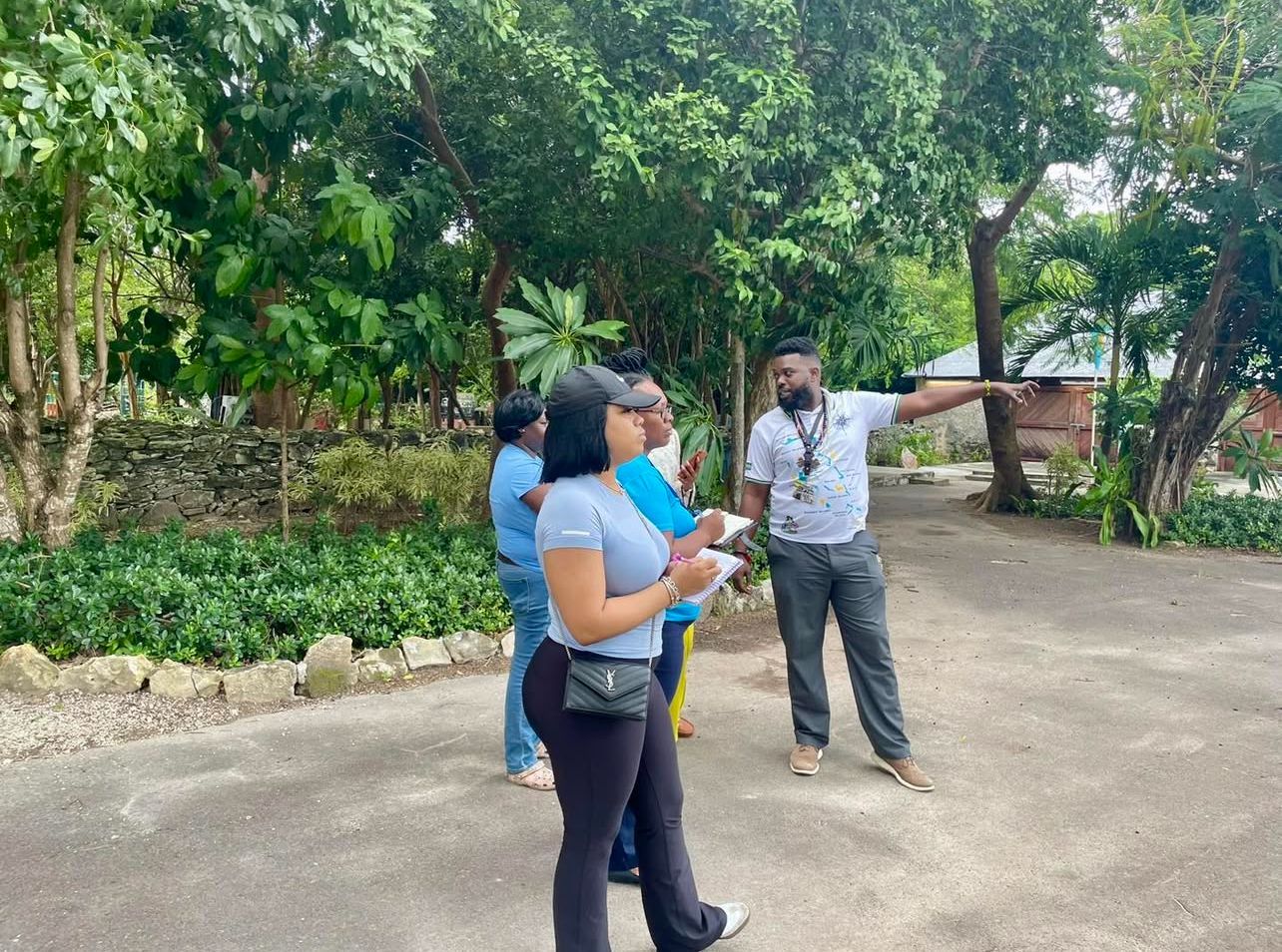 A group of people listens to a man pointing. They stand on a path surrounded by trees and greenery.