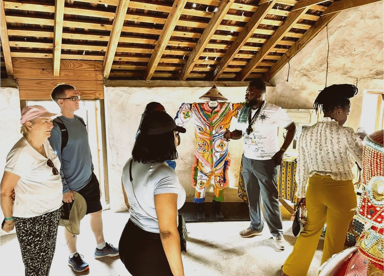 People listening to a guide, viewing artwork in a rustic building. Wooden ceiling, colorful clothing on display.