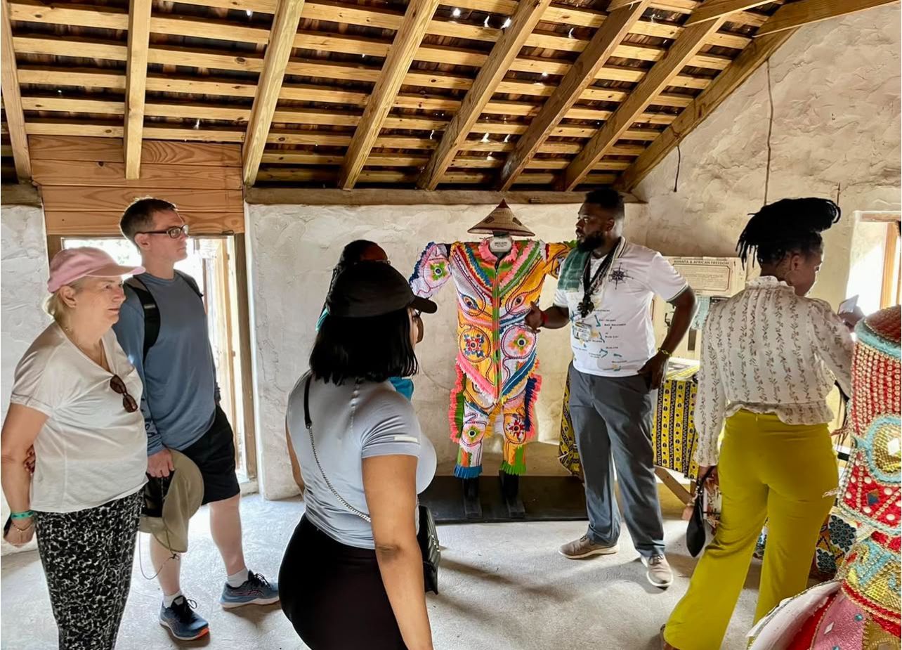 Tour group in a room looking at a colorful garment; wooden ceiling, white walls, and a speaker.