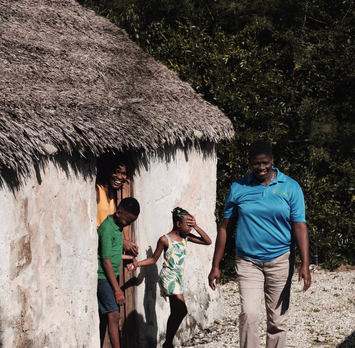 Family near a thatched-roof building. A woman walks, smiles, others look from doorway. Sunlight.