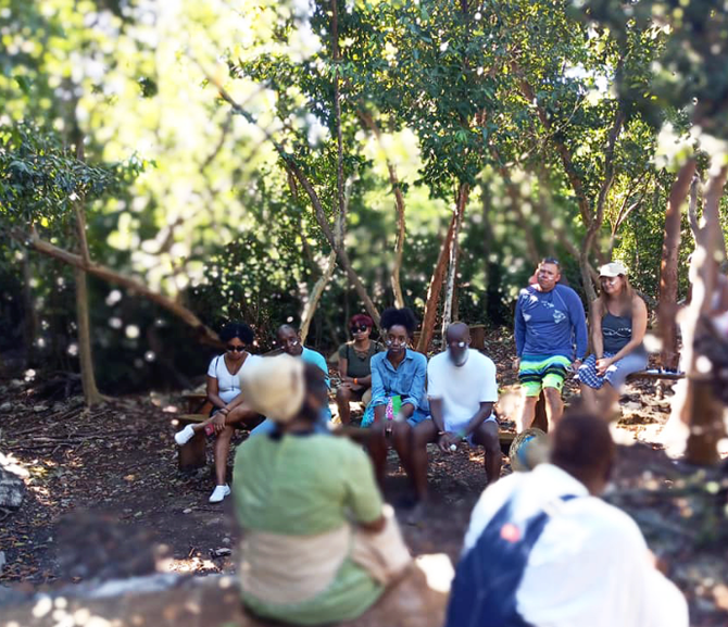 Group of people sitting on benches in a wooded area, listening to someone. Bright sunlight.