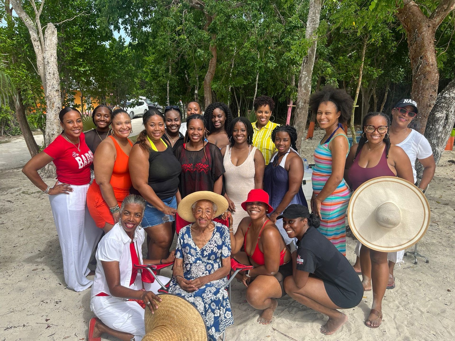 Group of women posing on a beach, smiling, and wearing summer attire. Trees and a car are in the background.