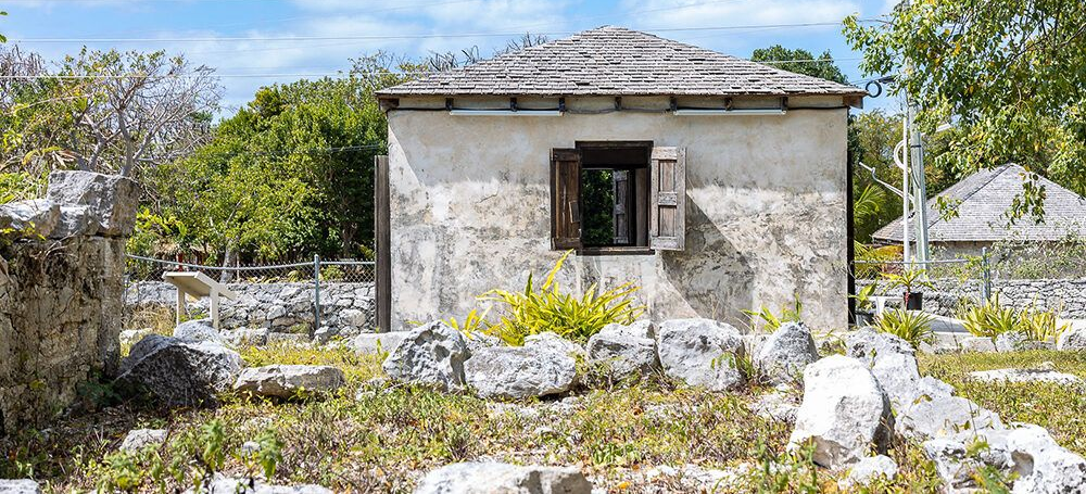 A small, weathered stone building with a wooden shuttered window sits in a rocky, overgrown area.