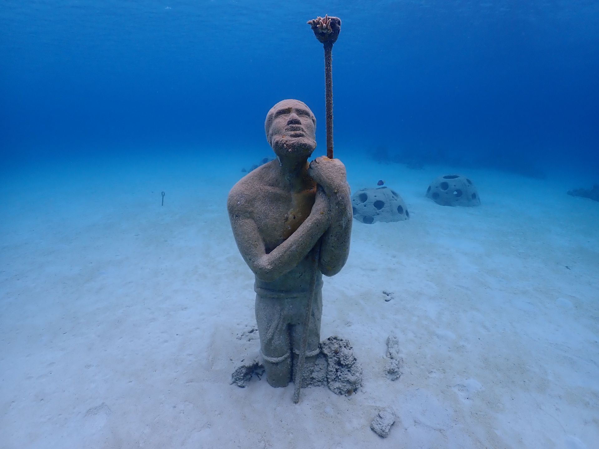 Underwater statue of a person holding a torch, sandy seabed, blue water.