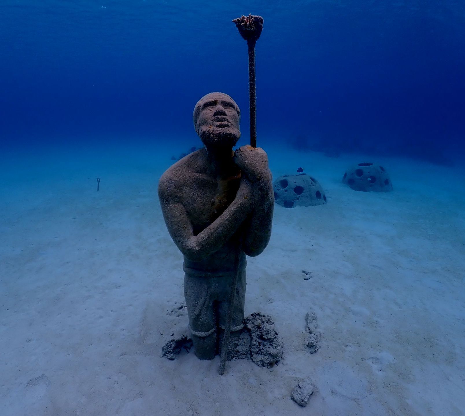 Underwater statue of a person holding a staff, sandy seabed, two coral-like structures in the background, deep blue water.