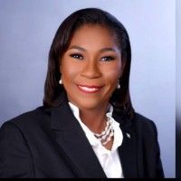 Woman in a dark suit smiles, wearing pearls and a white collared shirt, against a gray background.