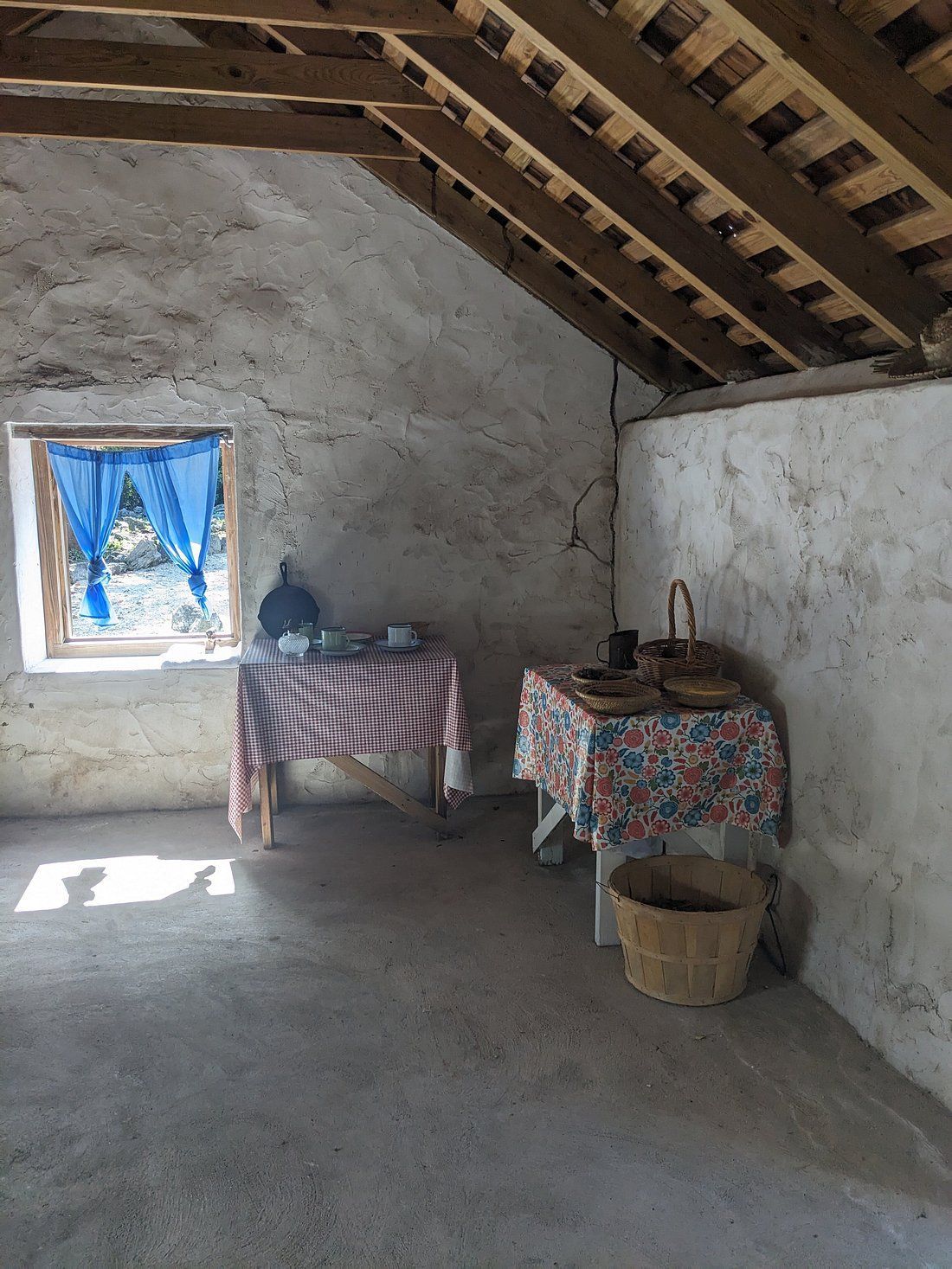 Interior of a rustic room with two tables covered in patterned cloths, a window with blue curtains, and a wooden basket.