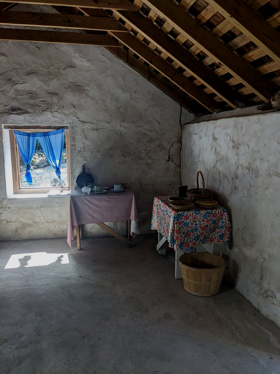 A rustic room with two tables covered in checkered and floral cloths, basket, and window with blue curtains.