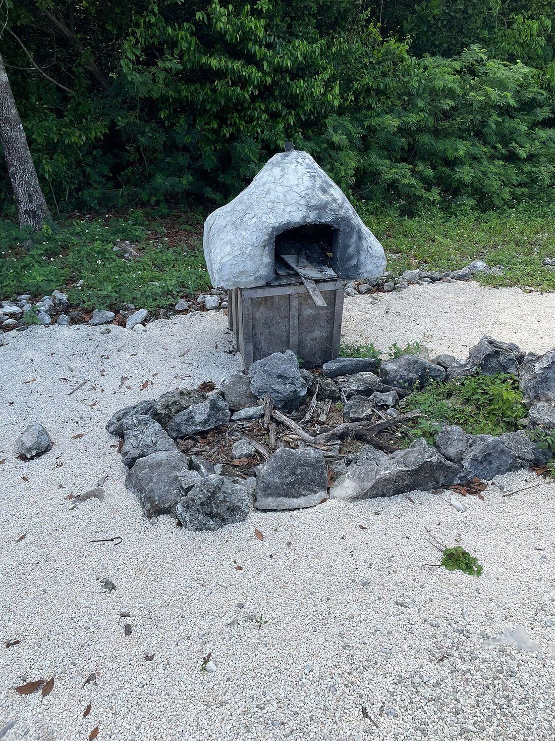 Outdoor stone oven structure on a gravel ground, surrounded by a ring of rocks. Green foliage in the background.