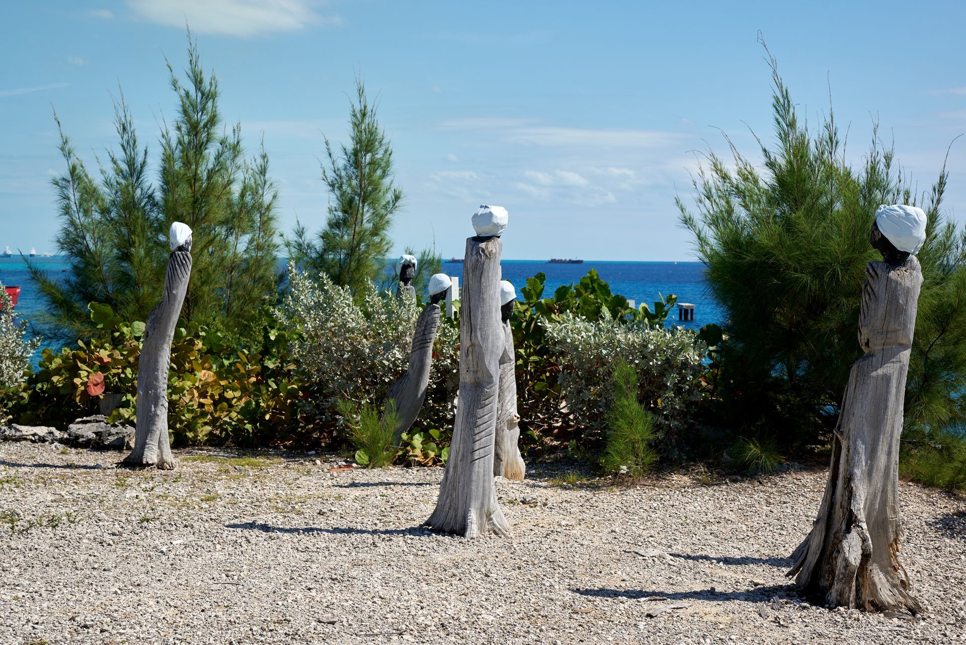 Sculptures on a pebble ground, with ocean in background, beneath a blue sky.