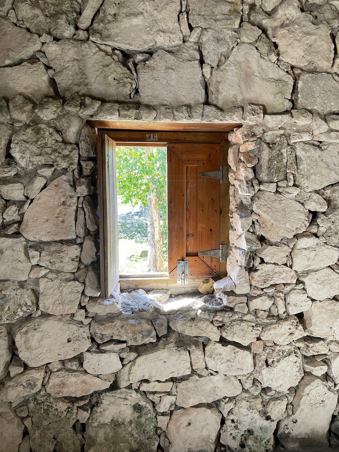 Stone wall with open wooden window frame; view of trees and sunlight.