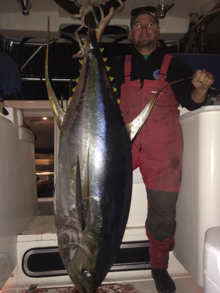 Man in red waders holds up large yellowfin tuna on a boat.