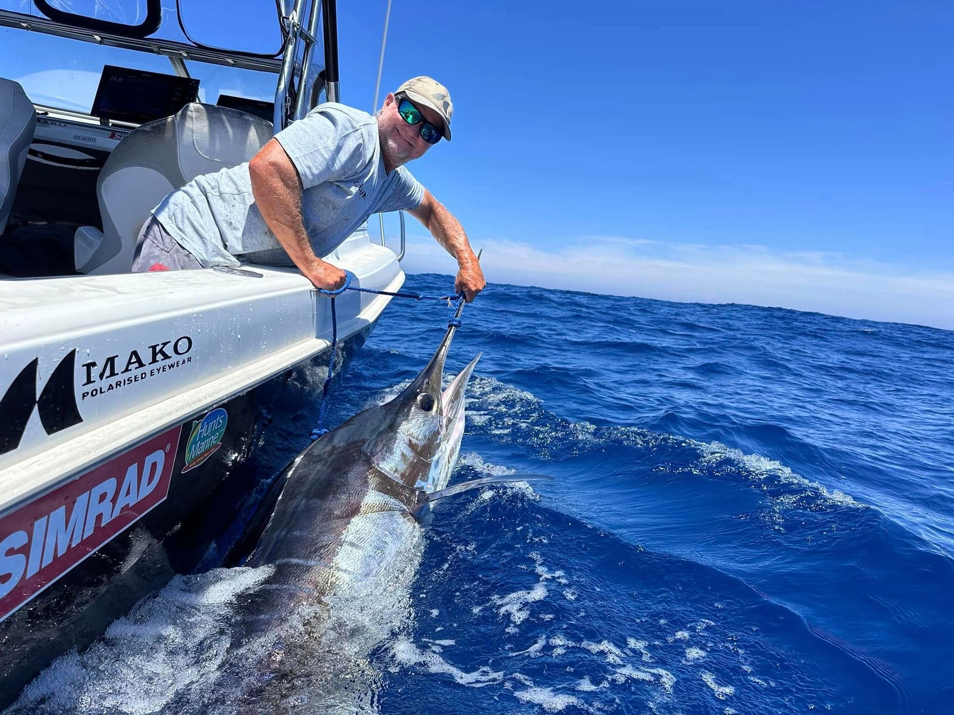 Man on boat, reeling in a large marlin in ocean, sunny day.
