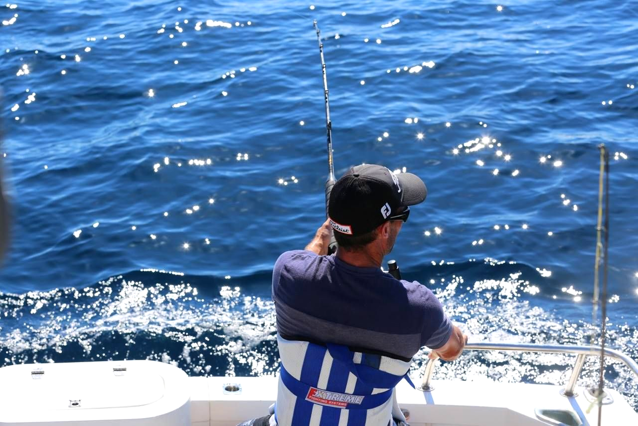 Man fishing on boat, holding rod. Blue water with sunlight reflecting.