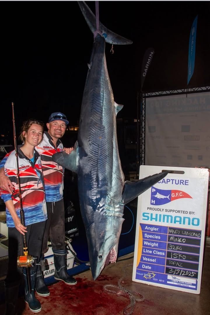 Man and girl pose with a large, hanging shark at night; a sign and blood are visible.