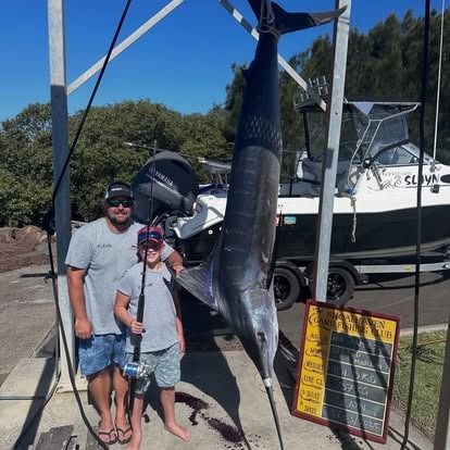 Father and child with a large marlin catch, hanging from a metal frame near a boat.