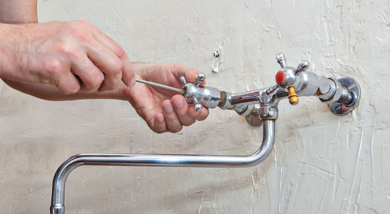 A person using a screwdriver to repair a chrome faucet mounted on a white wall.