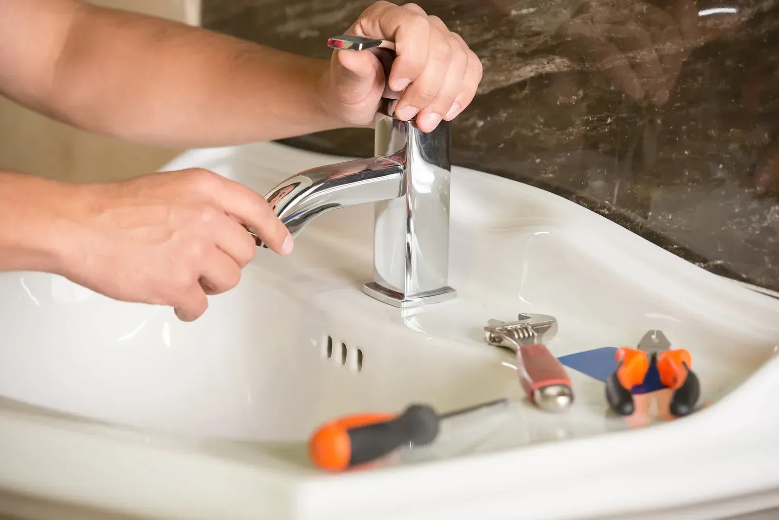 Hands repairing a chrome faucet in a bathroom sink, with tools lying nearby on the basin.