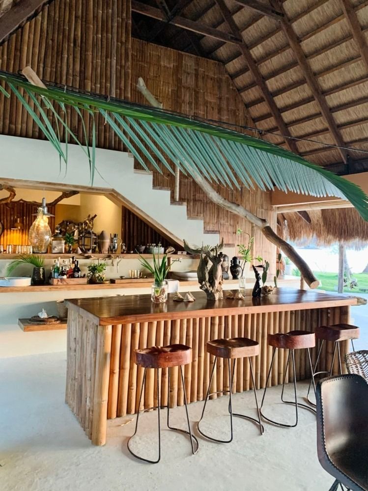 A kitchen with a wooden counter and stools and a thatched roof.