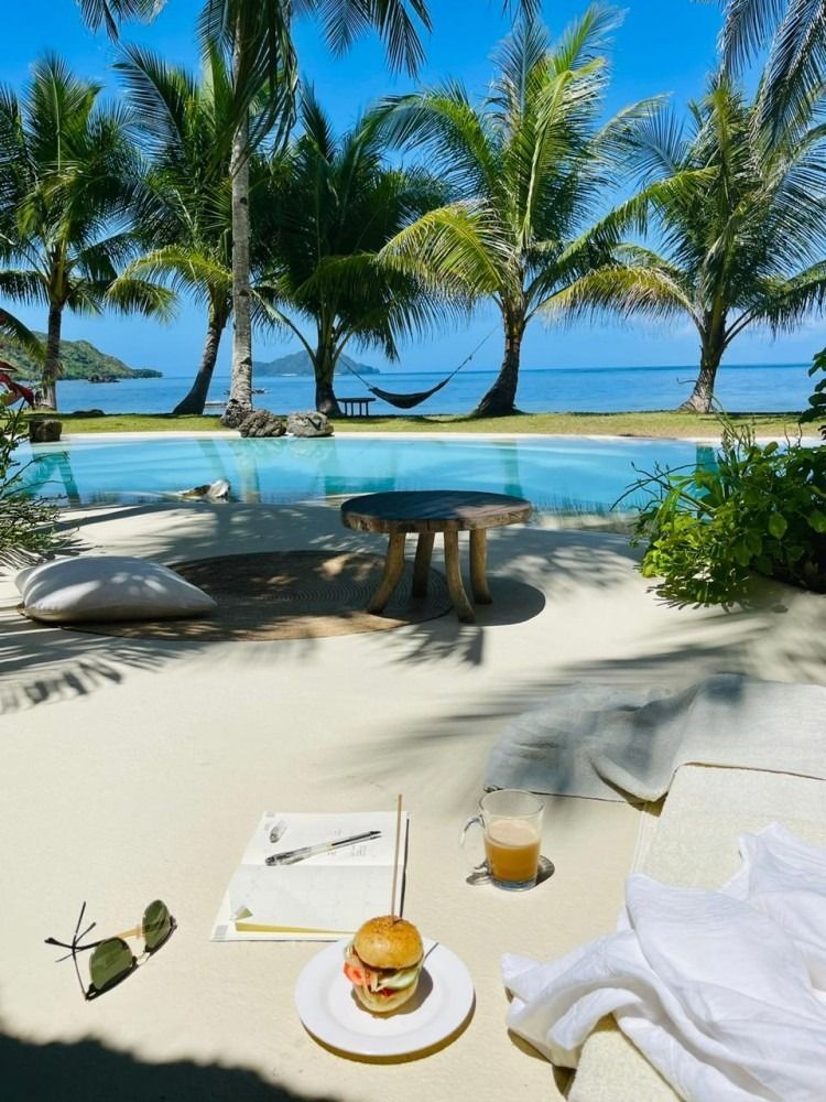 A hamburger is on a plate in front of a swimming pool surrounded by palm trees.