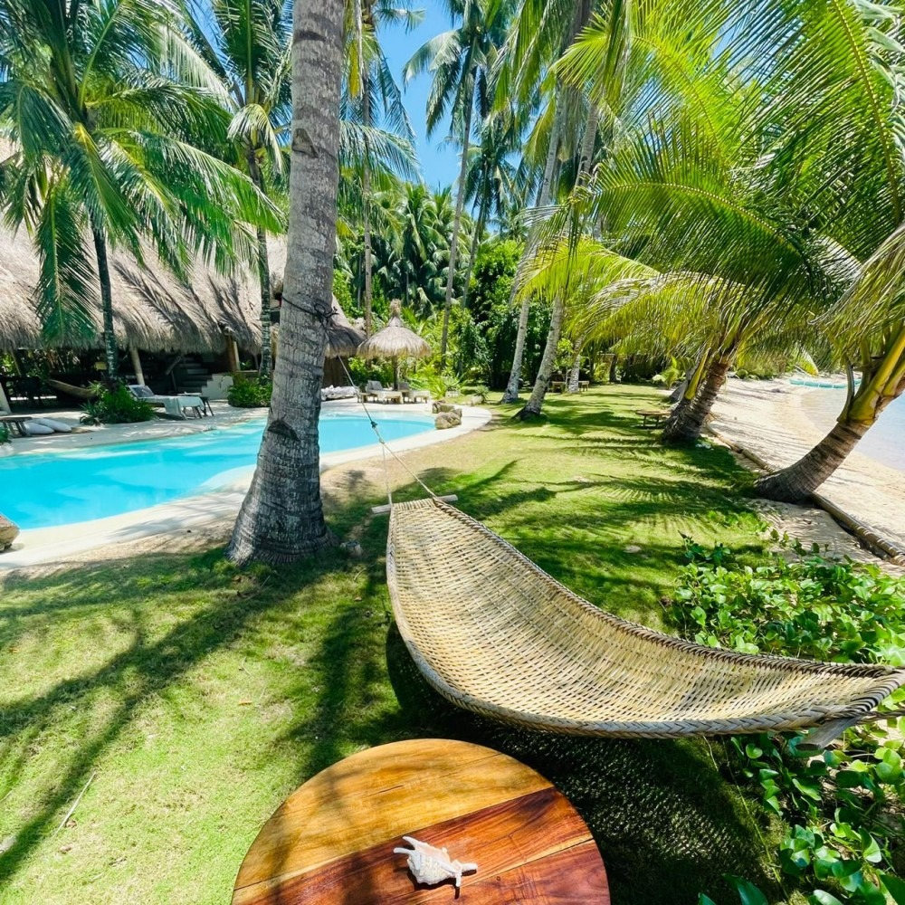 A hammock is sitting in front of a swimming pool surrounded by palm trees.