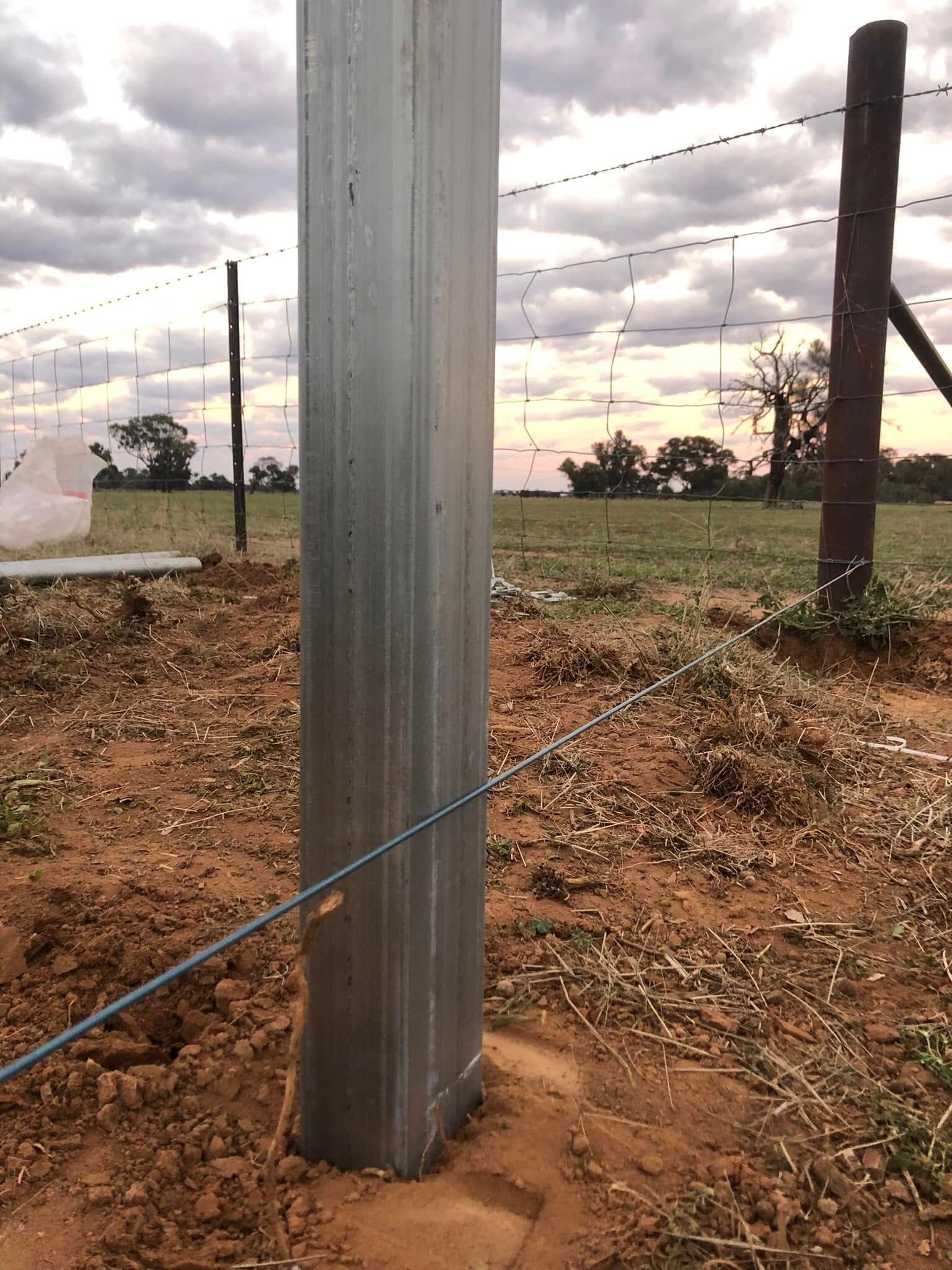 A metal pole is sitting in the middle of a dirt field next to a barbed wire fence.