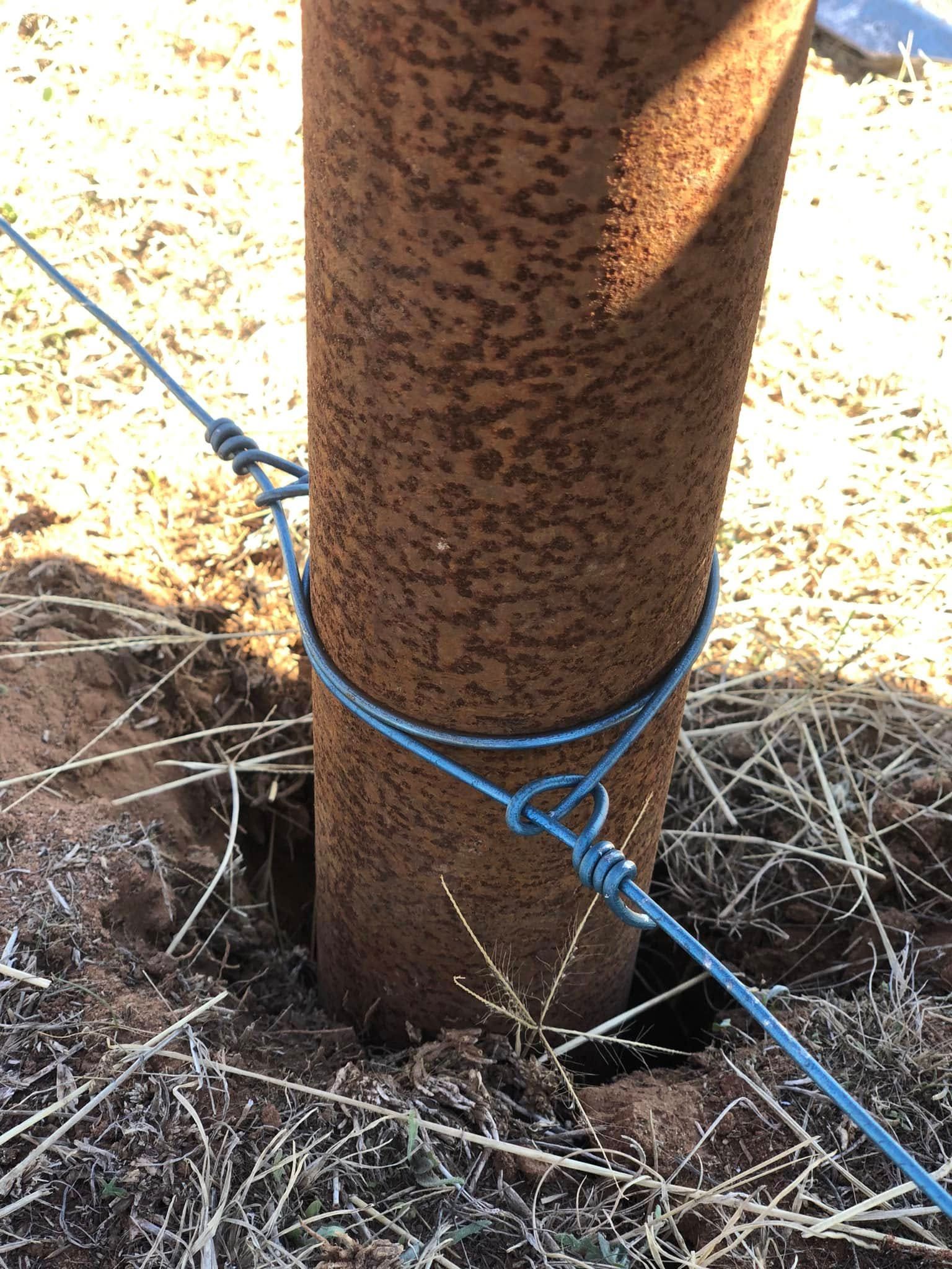 A rusty pipe is tied to a barbed wire fence post with a blue rope.