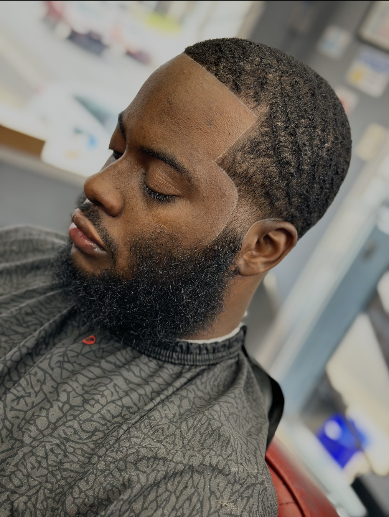 Man with a groomed beard and wavy haircut, wearing a barber's cape, eyes closed.