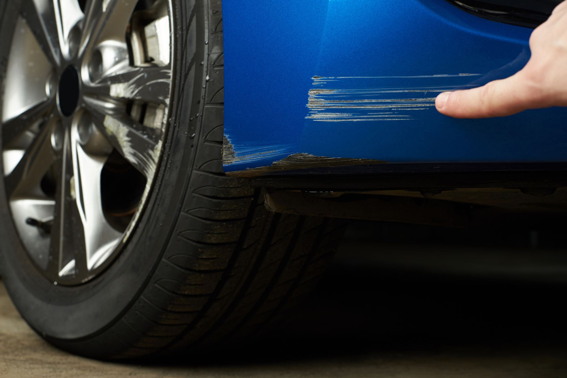 Scratched blue car bumper and tire close-up with hand pointing at damage for auto repair inspection.