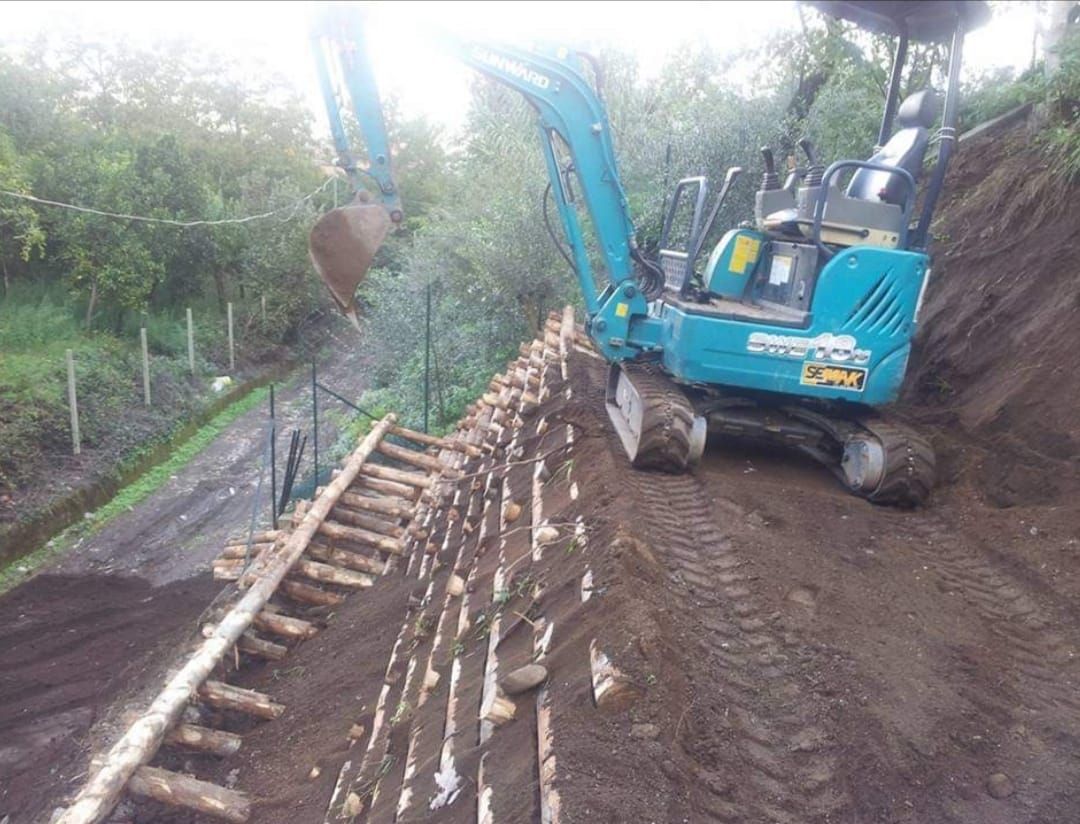 Escavatore blu che costruisce un muro di contenimento su una collina con travi di legno.