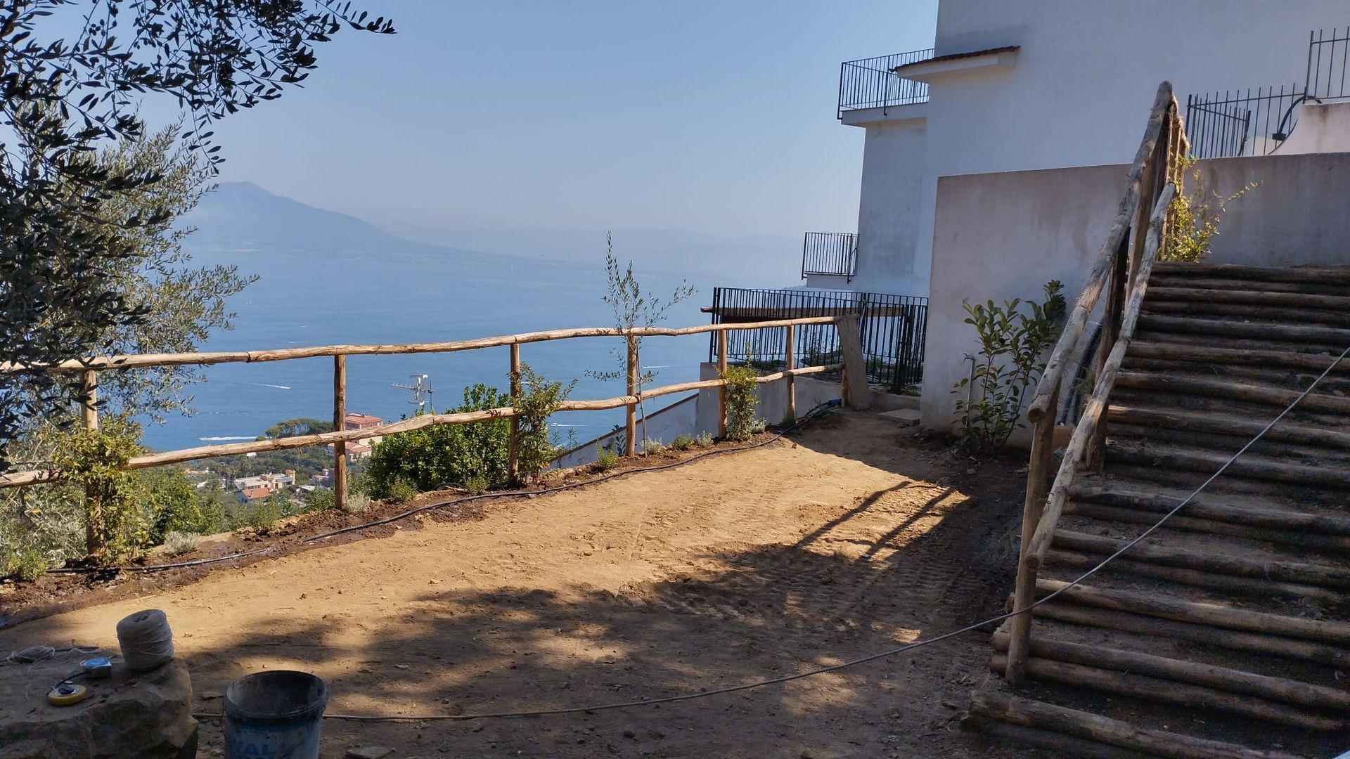 Una vista costiera con una staccionata in legno, un sentiero sterrato e gradini che conducono verso l'acqua e la montagna all'orizzonte.