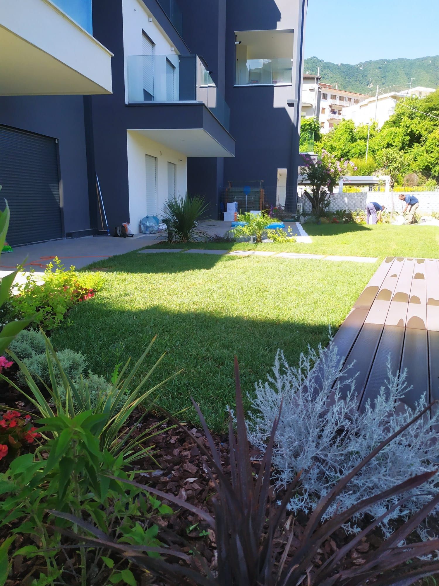 Un rigoglioso giardino verde con un edificio moderno sullo sfondo. Piante colorate delimitano il prato sotto un cielo luminoso e soleggiato.