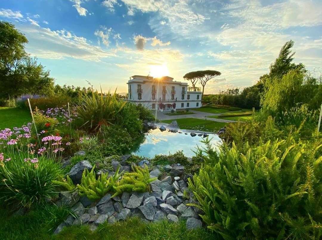 Villa soleggiata con laghetto e giardino. Un rigoglioso fogliame verde circonda l'acqua, con un cielo luminoso sullo sfondo.