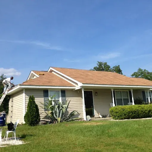 Person on ladder repairing a tan-roofed house with beige siding under a blue sky.