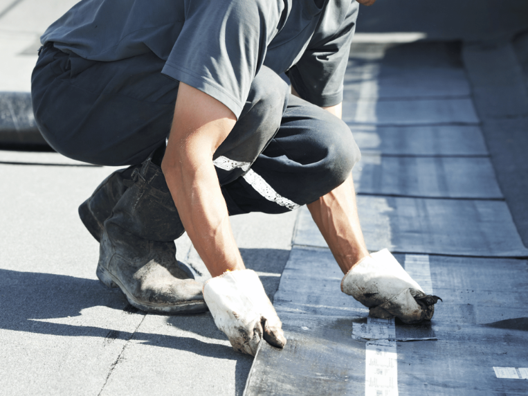 A construction worker in a yellow hard hat and gloves is installing roofing material on a flat roof.