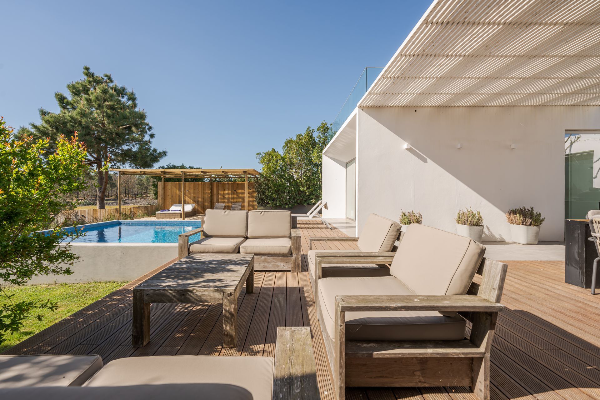 Modern patio with beige outdoor seating beside a white house and a swimming pool under a sunny sky