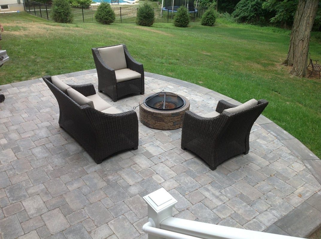 Outdoor patio with wicker chairs around a stone fire pit on a paver patio, with a grassy yard beyond.