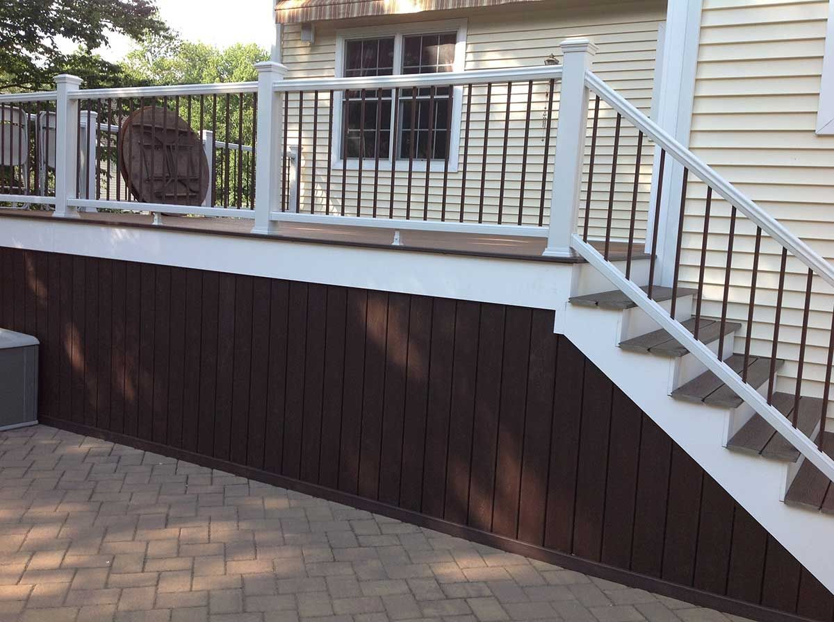 Backyard deck with white railing and stairs over dark retaining wall and brick patio