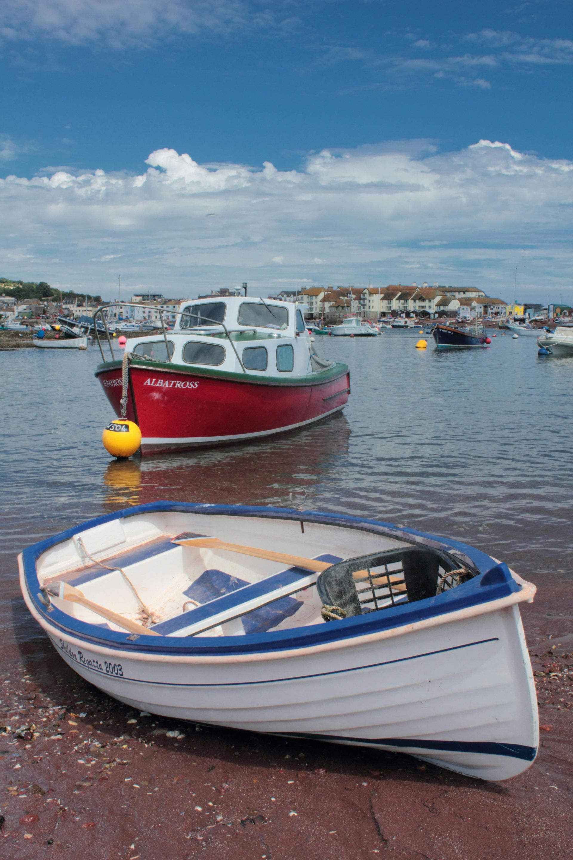 A small white boat on a red sandy shore, a red and white boat floats in water, buildings in the distance.